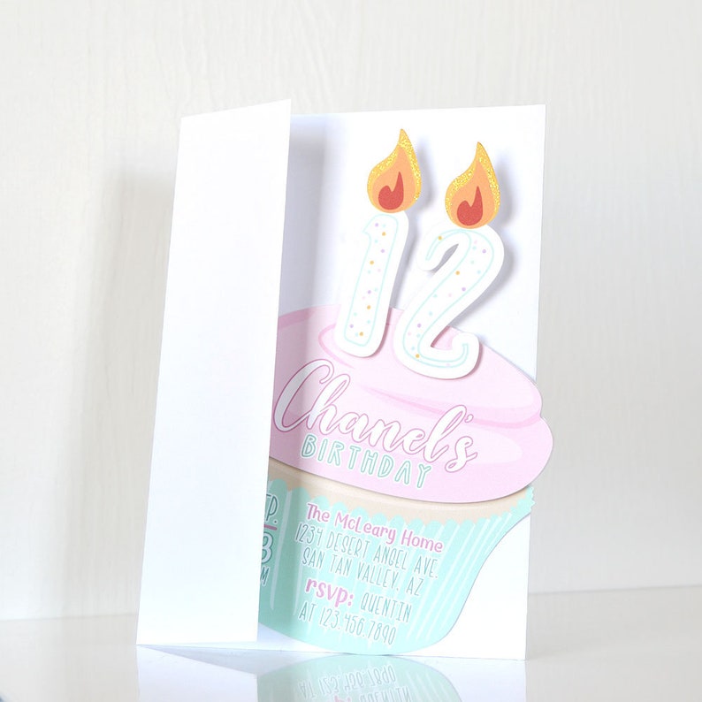 May include: A pink and blue cupcake with two lit candles that say "12" on top. The cupcake is on a white card with the text "Chanel's Birthday" and party details.