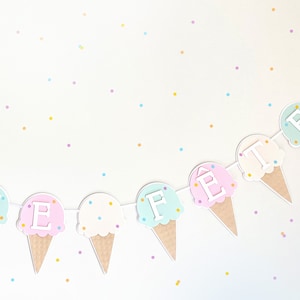 Op de afbeelding: Een pastelkleurige ijshoornbanner met de letters "FEte" erop. De hoorns zijn versierd met witte sprinkles en de letters zijn wit. De achtergrond is wit met pastelkleurig confetti.