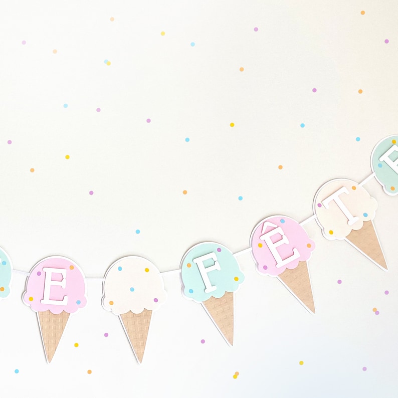 Op de afbeelding: Een pastelkleurige ijshoornbanner met de letters "FEte" erop. De hoorns zijn versierd met witte sprinkles en de letters zijn wit. De achtergrond is wit met pastelkleurig confetti.