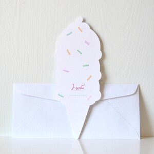 Puede incluir: Un sobre blanco con una tarjeta de cono de helado rosa, blanco y azul con chispas en la parte superior. La tarjeta dice "Lunita Papeleria" y "lunitapapeleria.com".