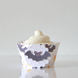 May include: A cupcake with white frosting in a Halloween-themed paper cup. The cup is white with a scalloped edge and features a repeating pattern of bats, dots, and candy corn.
