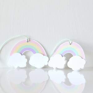 Pode incluir: Dois recortes de papel de arco-&iacute;ris pastel com nuvens brancas penduradas em um barbante. Os arco-&iacute;ris s&atilde;o em camadas de rosa, amarelo, verde e roxo.