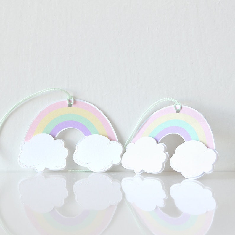 Pode incluir: Dois recortes de papel de arco-&iacute;ris pastel com nuvens brancas penduradas em um barbante. Os arco-&iacute;ris s&atilde;o em camadas de rosa, amarelo, verde e roxo.