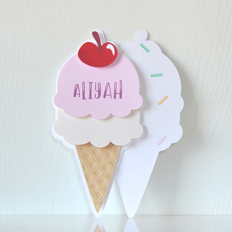 May include: A pink and white ice cream cone with a red cherry on top. The cone is decorated with a waffle pattern and the ice cream is topped with sprinkles. The cone says "Aliyah" on the side.