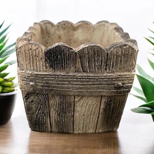 Op de afbeelding: Een vierkante, rustieke plantenbak van beton met een houtnerfdesign. De plantenbak heeft een gegolfde bovenrand en is geschilderd in tinten bruin en grijs.