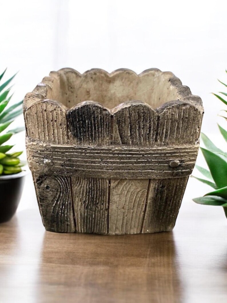 Op de afbeelding: Een vierkante, rustieke plantenbak van beton met een houtnerfdesign. De plantenbak heeft een gegolfde bovenrand en is geschilderd in tinten bruin en grijs.