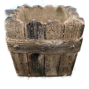 Op de afbeelding: Een kleine, vierkante plantenbak van bruin beton met een houtstructuur. De plantenbak heeft een gegolfde bovenrand en een verhoogde rand rond de bovenkant.