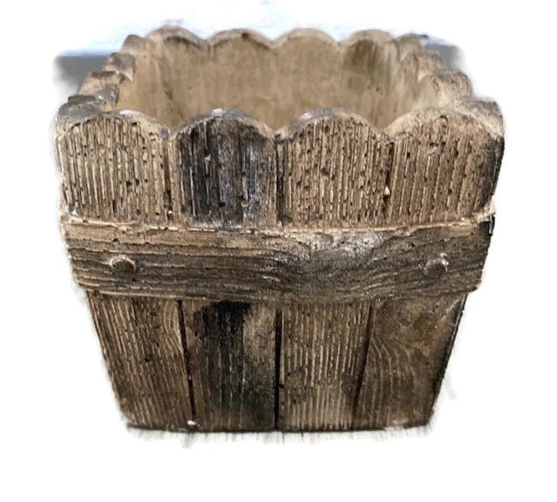 Op de afbeelding: Een kleine, vierkante plantenbak van bruin beton met een houtstructuur. De plantenbak heeft een gegolfde bovenrand en een verhoogde rand rond de bovenkant.