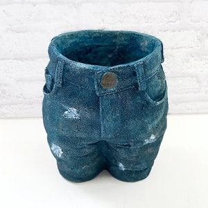 May include: A blue denim-style planter pot. The pot is shaped like a pair of jeans, complete with pockets, belt loops, and a button. The denim has distressed details with white paint accents. The pot is designed to hold small plants or flowers.