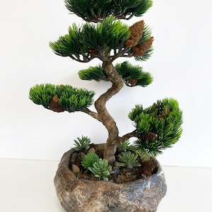 May include: A green artificial bonsai tree with brown pine cones in a brown and gray rock-shaped pot. The tree has a twisted trunk and multiple branches with green leaves.