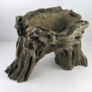May include: A decorative planter with a rustic, weathered appearance. The planter is designed to resemble a section of a tree trunk, with intricate details of bark and wood grain. The color is a blend of gray and brown, creating a natural, earthy aesthetic.