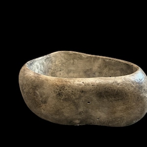 May include: A bowl with an irregular, organic shape and a textured, weathered surface in shades of gray and brown. The interior is smooth and light gray, contrasting with the rough, stone-like exterior finish of this decorative vessel.