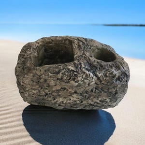 May include: A gray, textured rock with two indentations, resembling a planter, on a white sandy beach.
