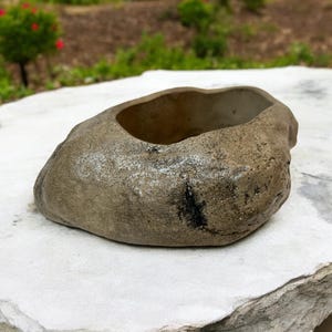May include: A gray stone planter with a rough, textured surface. The planter is shaped like a rounded bowl and has a wide opening.