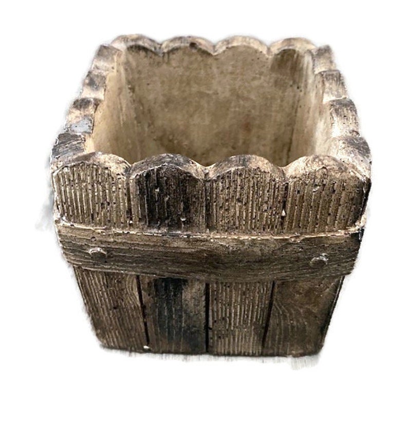 Op de afbeelding: Een vierkante plantenbak van beton met een houtlook. De plantenbak heeft een gegolfde bovenrand en een rustieke uitstraling.