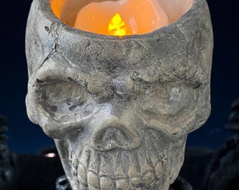 May include: A gray, textured skull candle holder with a flickering LED candle inside. The skull has a detailed face with a wide mouth and large eyes.