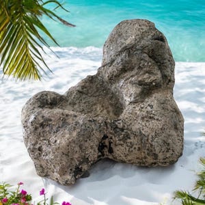 May include: A large, gray, textured rock with a rounded shape sits on a white sandy beach. The rock is smooth and has a natural, weathered look.