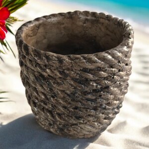 May include: A gray concrete planter with a rope-like texture. The planter is empty and sitting on a white sandy beach.