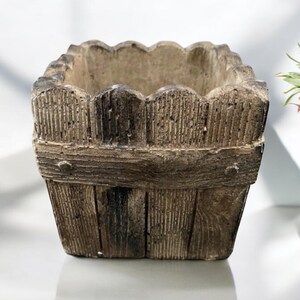 Op de afbeelding: Een vierkante, bruine betonnen plantenbak met een houtstructuur en een gegolfde bovenrand. De plantenbak is ontworpen om eruit te zien als een houten hek.
