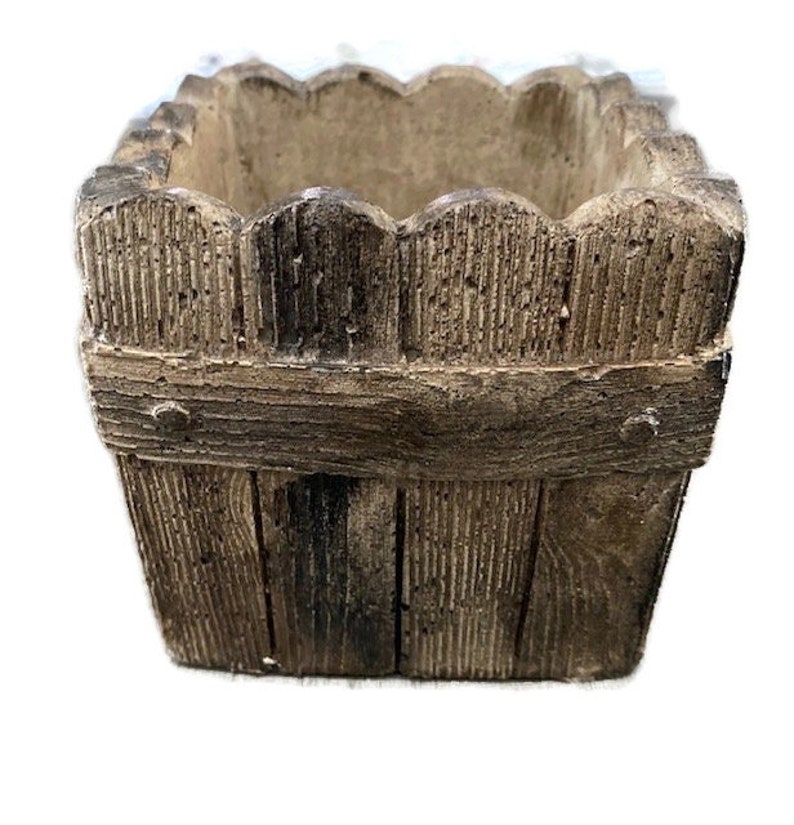 Op de afbeelding: Een vierkante plantenbak gemaakt van bruin beton met een houtstructuur. De plantenbak heeft een verhoogde rand en een licht gegolfde rand. Hij is ontworpen om eruit te zien als een houten kist.