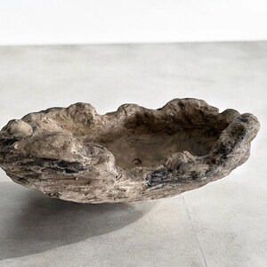 May include: A decorative, oval-shaped bowl with a rough, textured surface. The bowl is primarily beige with dark gray and black accents, giving it a natural, weathered appearance. The rim has an uneven, organic shape.