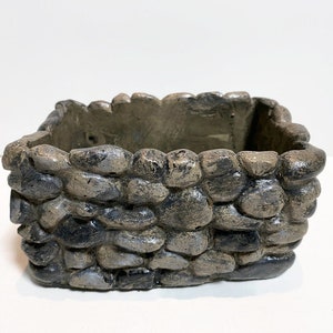 May include: A small, square planter made of gray concrete with a rough, stone-like texture. The planter is designed to look like a miniature stone wall.
