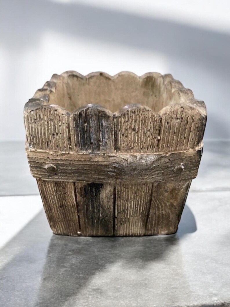 Op de afbeelding: Een vierkante betonnen plantenbak met een houtlook. De plantenbak heeft een gegolfde bovenrand en een rustieke bruine kleur.