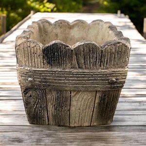 Op de afbeelding: Een vierkante betonnen plantenbak met een houtnerfdesign. De plantenbak heeft een gegolfde bovenrand en is gemaakt om eruit te zien als een houten vat.