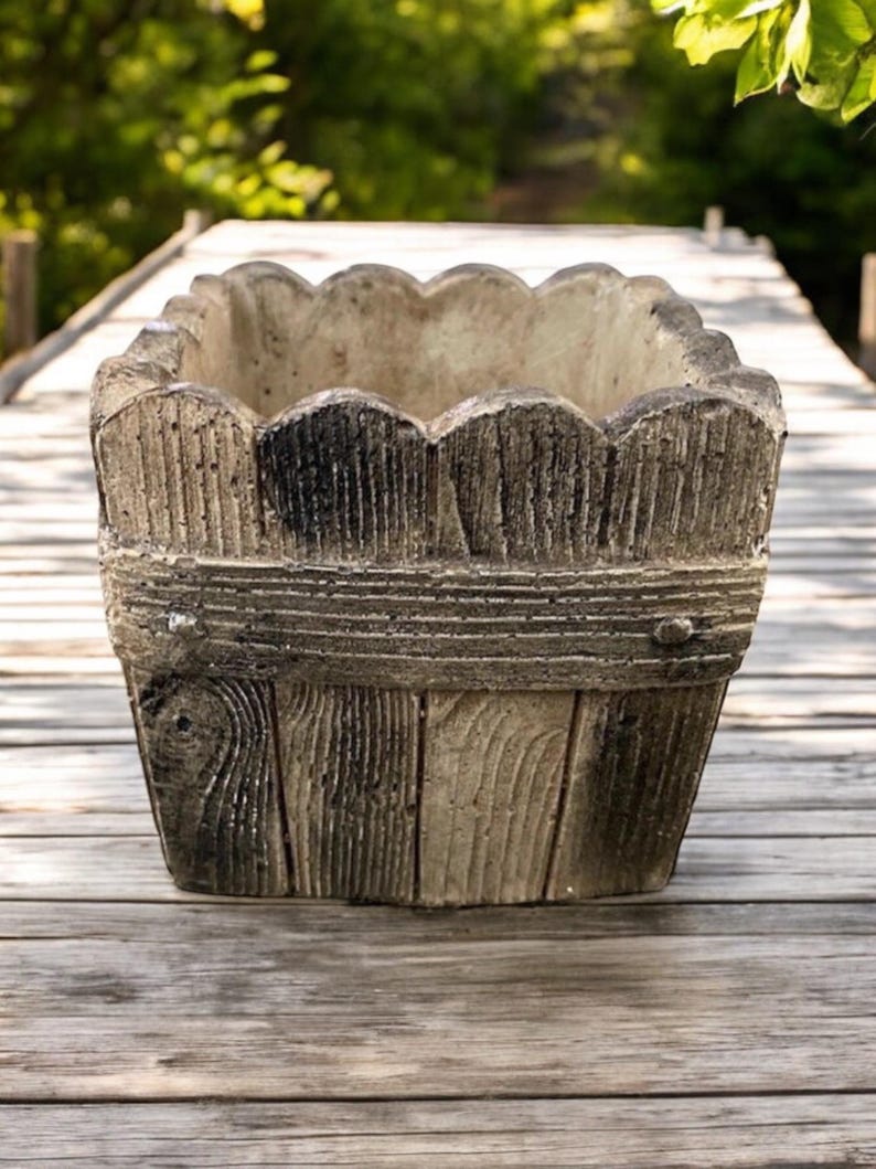Op de afbeelding: Een vierkante betonnen plantenbak met een houtnerfdesign. De plantenbak heeft een gegolfde bovenrand en is gemaakt om eruit te zien als een houten vat.
