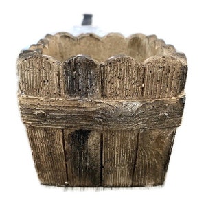 Op de afbeelding: Een vierkante, bruine, houten plantenbak met een decoratief hekwerkontwerp rond de bovenrand.