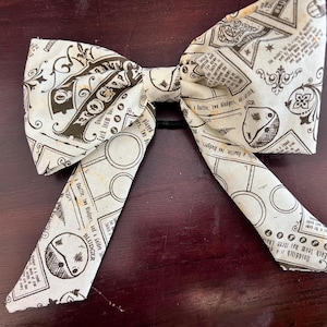 May include: A cream-colored bow tie with a black print of text and illustrations. The bow tie is tied in the center with a black elastic band. The fabric has a vintage, aged appearance.