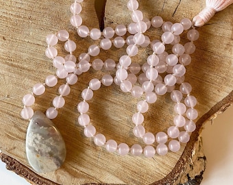 May include: A mala necklace featuring smooth, round, pale pink rose quartz beads strung on a cord, finished with a light pink tassel and a large, teardrop-shaped pendant made of a mottled gray and white stone with subtle pink inclusions.