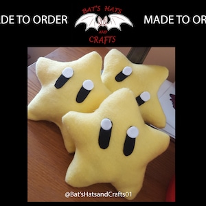 Peut inclure: Trois peluches jaunes en forme d'étoile avec des yeux noirs et des pupilles blanches. Les peluches sont sur une surface en bois. Le texte "Bat's Hats and Crafts" est visible en arrière-plan.