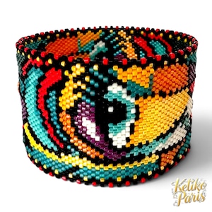 Könnte beinhalten: Ein breites Armband aus kleinen Glasperlen in einem lebendigen, mehrfarbigen abstrakten Muster. Das Design zeigt kräftige Wirbel in Gelb, Türkis, Rot, Lila und Orange, die alle mit schwarzen Perlen umrandet sind. Der Text Ketiko Paris ist sichtbar.