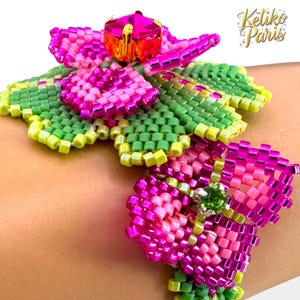 Peut inclure: Un bracelet de perles avec un grand motif floral stylisé. Les pétales de la fleur sont d'un rose vif, avec des accents verts et jaunes. Une plus petite fleur est également visible. Le bracelet est orné d'une gemme rose et du texte "Ketiko Paris".