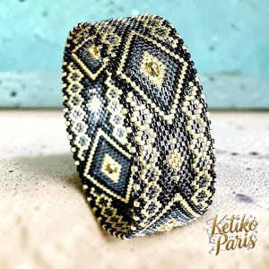 Peut inclure: Un bracelet manchette large composé de petites perles de verre avec un motif géométrique noir, or métallique et gris. Le design présente des losanges répétitifs et des détails en treillis. Le texte Ketiko Paris est visible dans le coin.