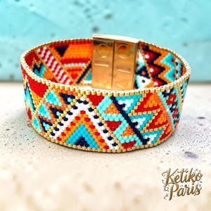 Peut inclure: Un large bracelet en perles avec un motif géométrique de triangles et de zigzags dans des tons vifs d'orange, turquoise, rouge, blanc et bleu foncé. Le bracelet comprend un fermoir magnétique rectangulaire doré et de petites perles dorées.