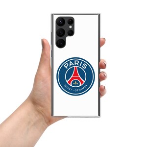 Puede incluir: Una funda transparente para teléfono con el logotipo del Paris Saint-Germain. El logotipo es un emblema circular con un borde azul, la Torre Eiffel en rojo y las palabras "PARIS" y "SAINT-GERMAIN". La funda está en una mano.