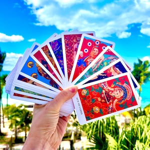May include: A deck of tarot cards fanned out against a blue sky with clouds. The cards feature colorful illustrations with various figures and symbols. The Ace of Coins card is visible, with red roses and the card's title in white text.