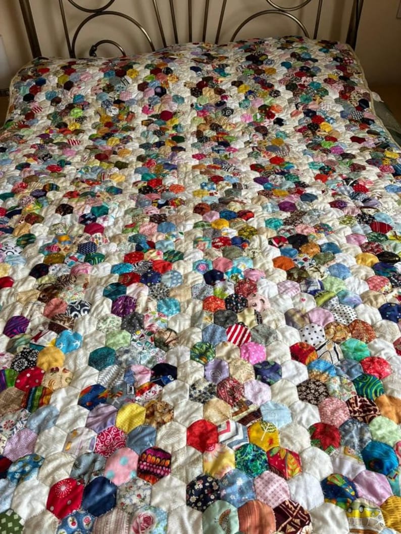 May include: A colorful quilt with a honeycomb pattern. The quilt is made of many small, hexagonal fabric pieces in a variety of colors and patterns, including floral, striped, and solid fabrics. The quilt is on a bed with a metal headboard.