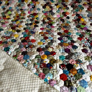 May include: A handmade quilt featuring a patchwork design of colorful, hexagonal fabric pieces. The quilt showcases a variety of patterns and colors, including red, blue, green, and yellow, set against a white background. The quilt is on a bed with a white textured blanket.