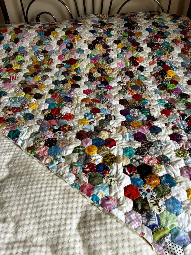 May include: A handmade quilt featuring a patchwork design of colorful, hexagonal fabric pieces. The quilt showcases a variety of patterns and colors, including red, blue, green, and yellow, set against a white background. The quilt is on a bed with a white textured blanket.