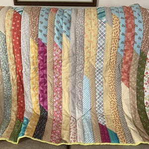 May include: A patchwork quilt with vertical strips of patterned fabrics in various colours. The quilt features floral, geometric, and solid designs in yellow, blue, pink, and green. The quilt has a yellow border around the edge.