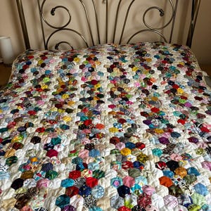 May include: A colourful patchwork quilt with a hexagonal design, featuring a variety of fabric patterns and colours. The quilt, with red, blue, yellow, and green hues, is set against a white backdrop. It is displayed on a bed with a decorative metal headboard.