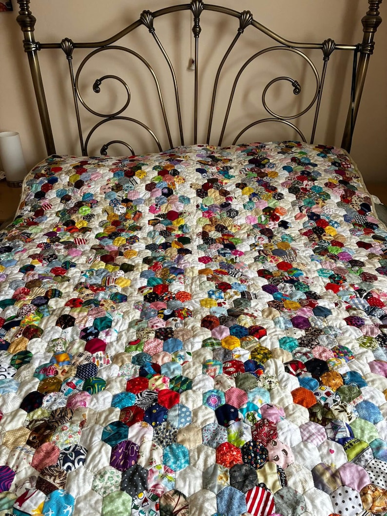 May include: A handmade quilt with a patchwork design of colorful hexagonal fabric pieces. The quilt features a variety of patterns and colors, including red, blue, yellow, and green, set against a white background. The quilt is draped over a bed with a decorative metal headboard.