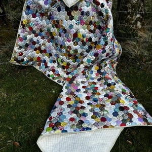 May include: A handmade quilt with a patchwork design of colorful hexagonal fabric pieces. The quilt features a white, textured backing and a white border. The quilt is draped over a grassy surface, showcasing its vibrant colors and intricate pattern.