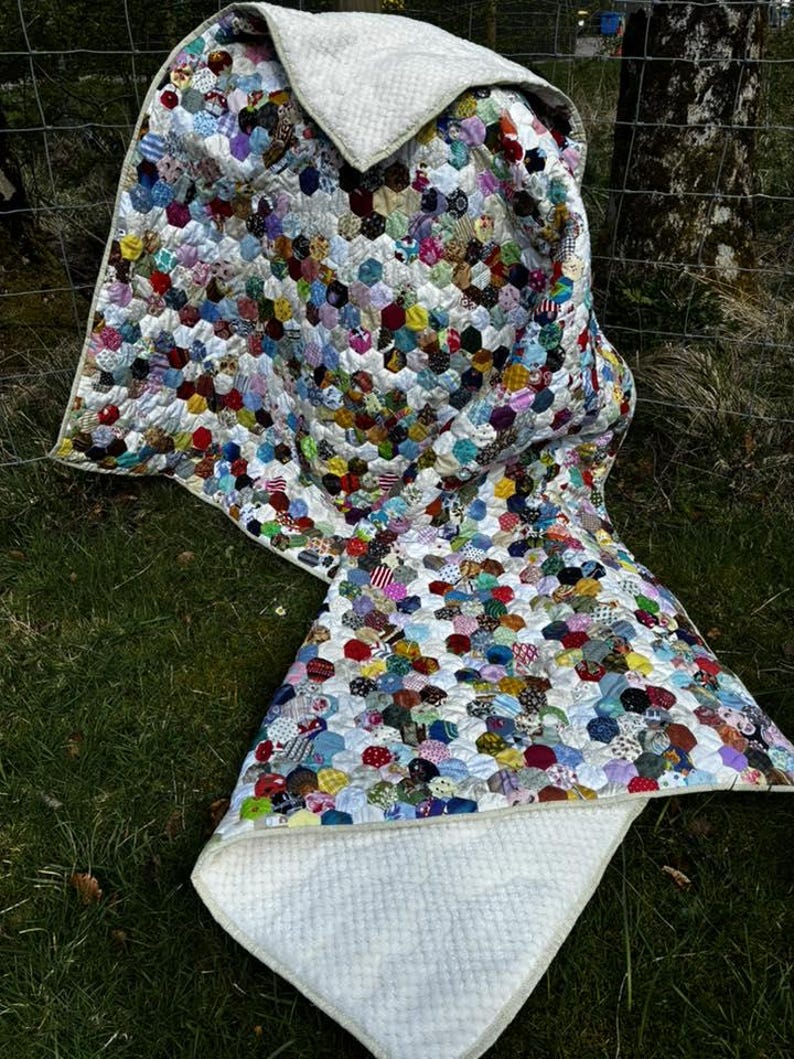 May include: A handmade quilt with a patchwork design of colorful hexagonal fabric pieces. The quilt features a white, textured backing and a white border. The quilt is draped over a grassy surface, showcasing its vibrant colors and intricate pattern.