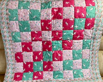 May include: A handmade quilt featuring a patchwork design with squares of pink, teal, and fuchsia fabric. Each square showcases a white unicorn print. The quilt is bordered with a patterned fabric and is likely for a child's bed.