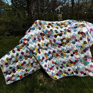 May include: A handmade quilt with a white background and colorful, patterned hexagonal patches. The quilt is spread out on green grass, with a wooden fence and trees in the background. The patches are in various colors, including red, blue, yellow, and green.