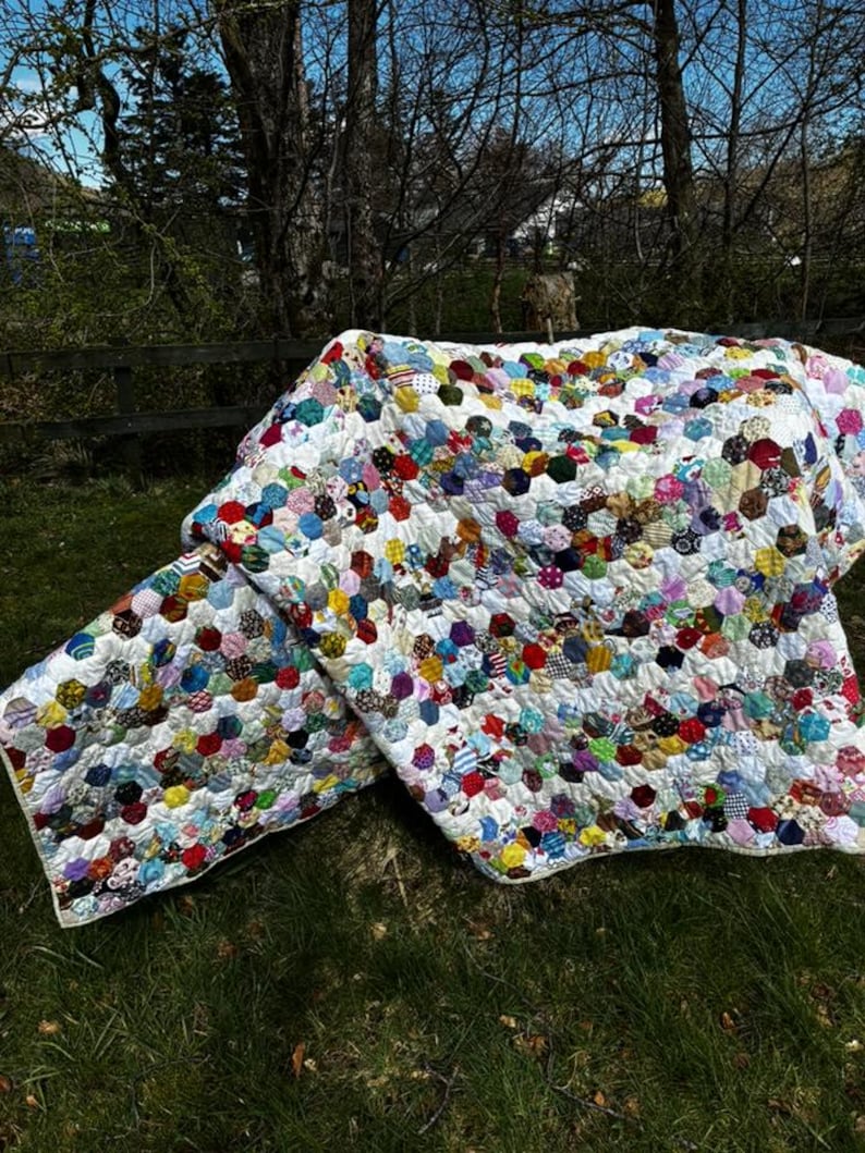 May include: A handmade quilt with a white background and colorful, patterned hexagonal patches. The quilt is spread out on green grass, with a wooden fence and trees in the background. The patches are in various colors, including red, blue, yellow, and green.
